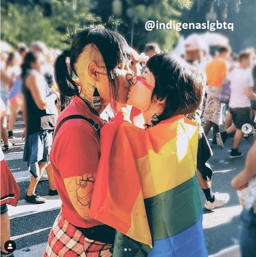 Duas pessoas indígenas se beijando, envoltas em uma bandeira do arco-íris