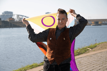 Pessoa sorrindo à beira de um rio, segurando a bandeira intersexo aberta atrás de si